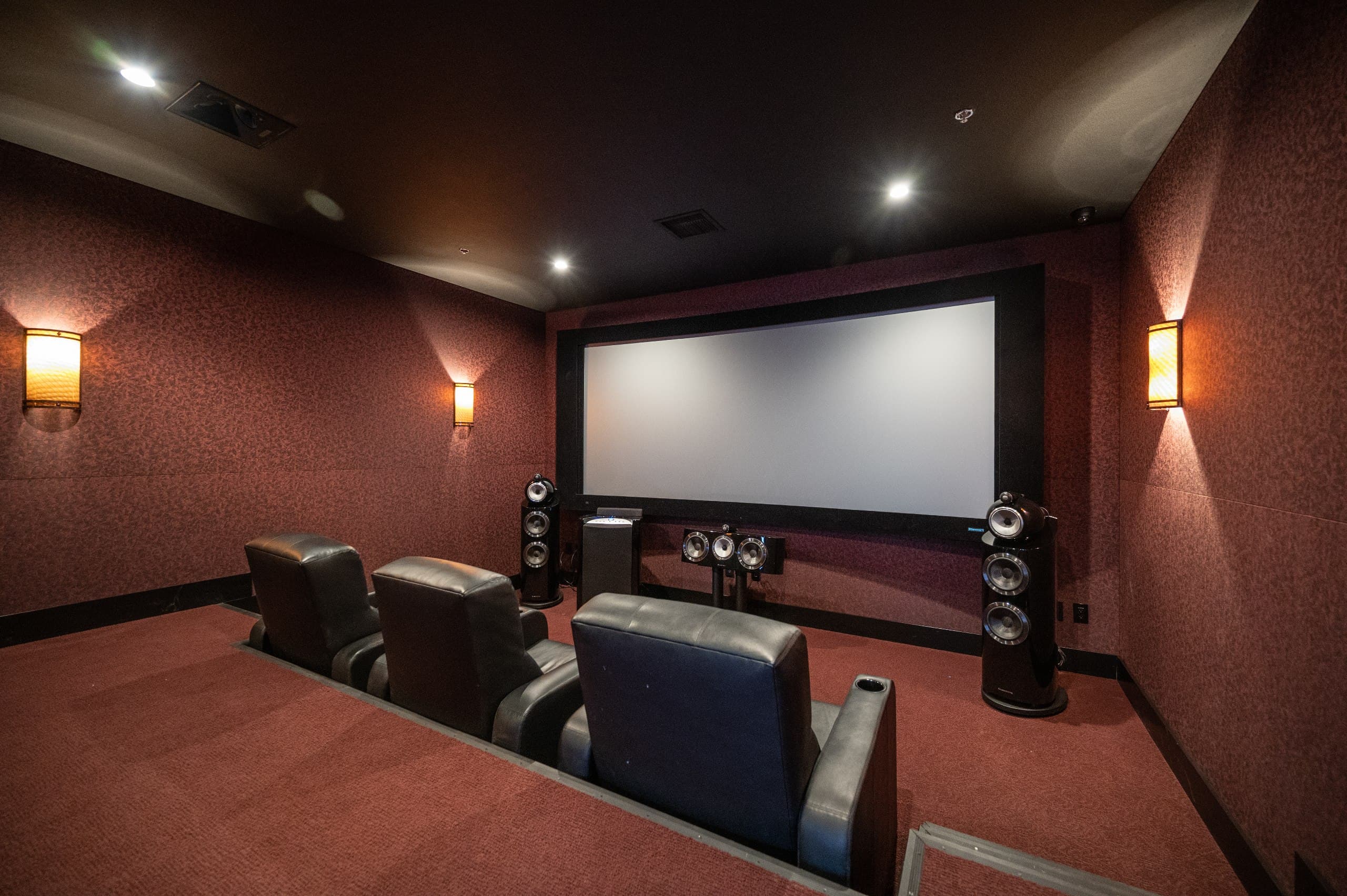 Dedicated cinema room with floor-standing B&W speakers flanking a large projection screen with sconce lighting